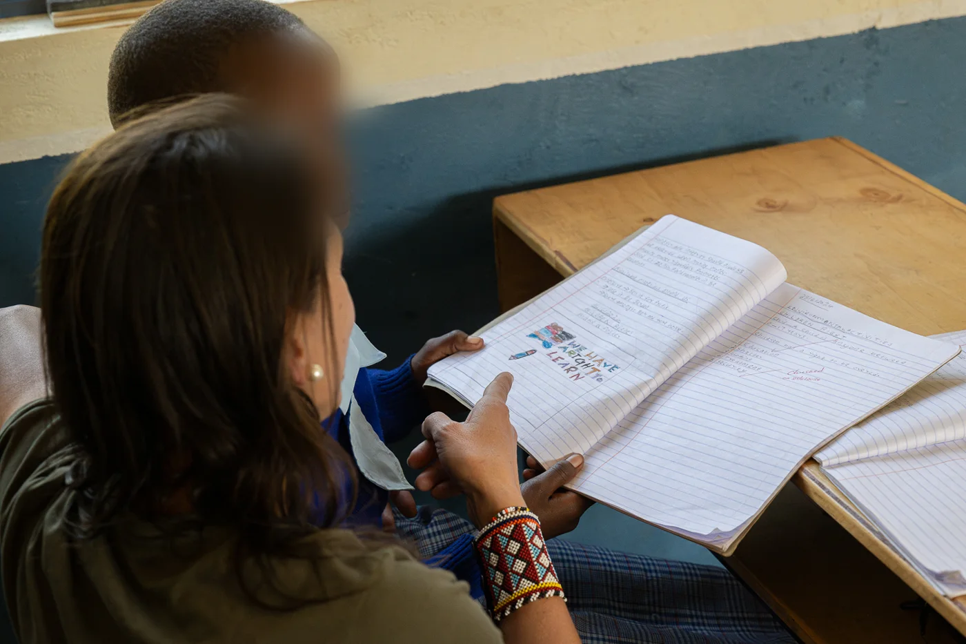 Voluntaria revisando con un alumno su cuaderno, con el lema «We have a right to learn» (rostro del menor difuminado)